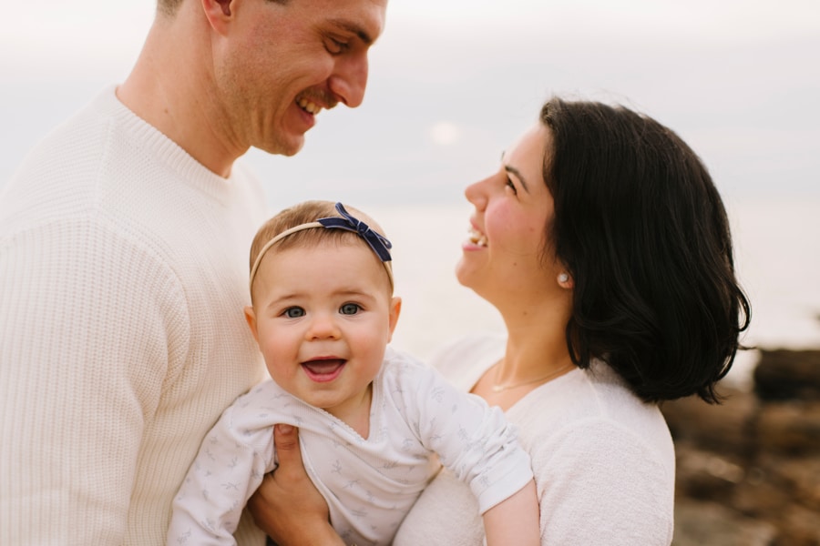 Andrea Bombino and her husband smile at each other as their baby smiles at the camera.